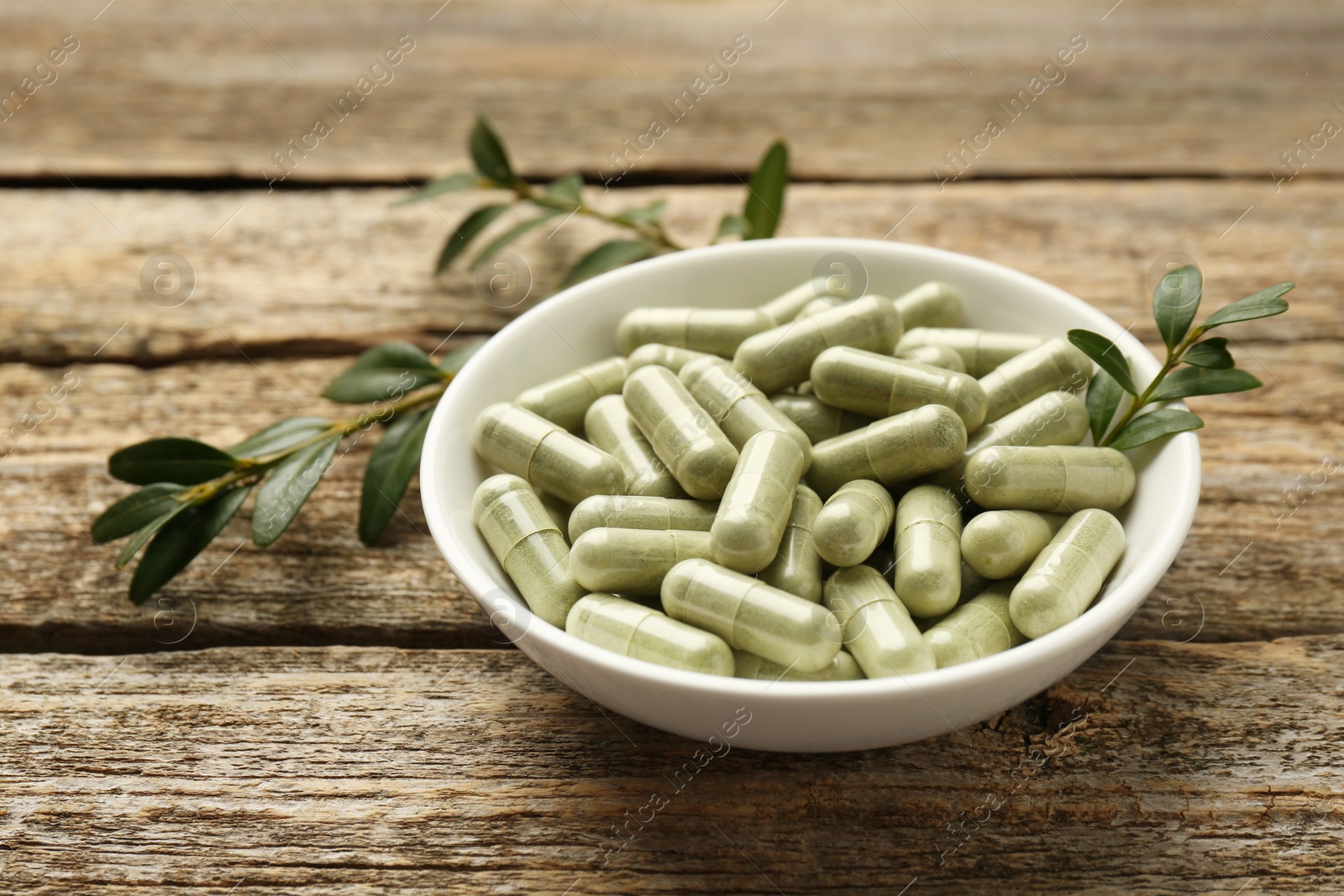 Photo of Vitamin capsules in bowl and leaves on wooden table, closeup