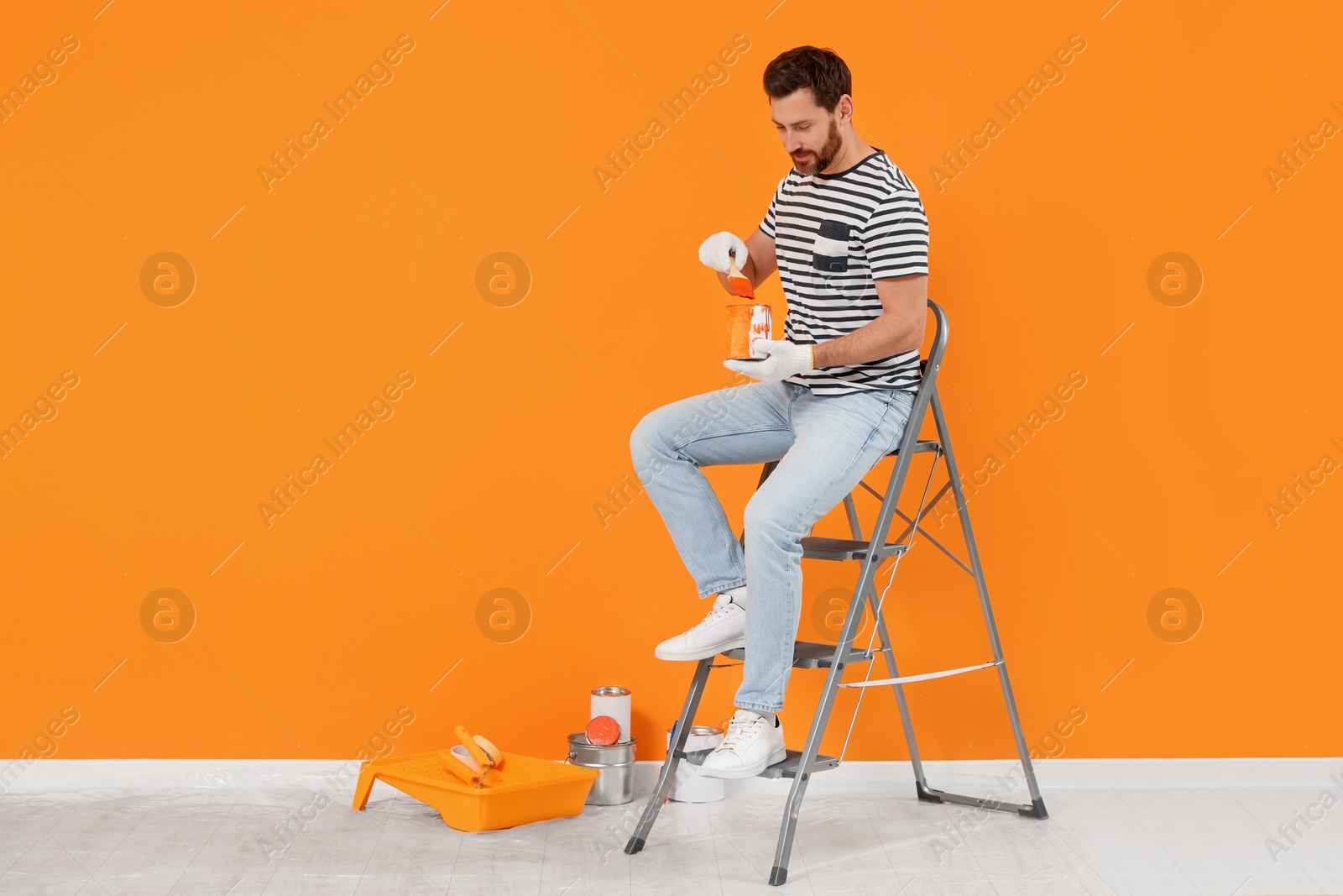 Photo of Designer with painting equipment sitting on folding ladder near freshly painted orange wall indoors, space for text