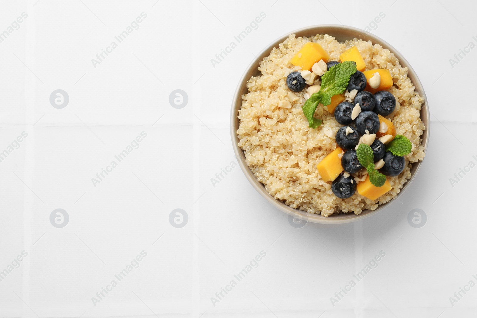 Photo of Tasty quinoa porridge with blueberries, pumpkin and mint in bowl on white tiled table, top view. Space for text