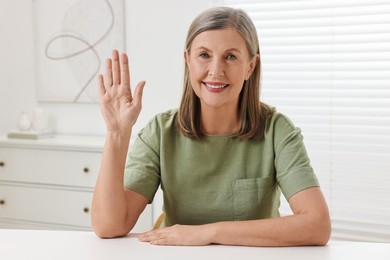 Happy woman waving hello at white table indoors