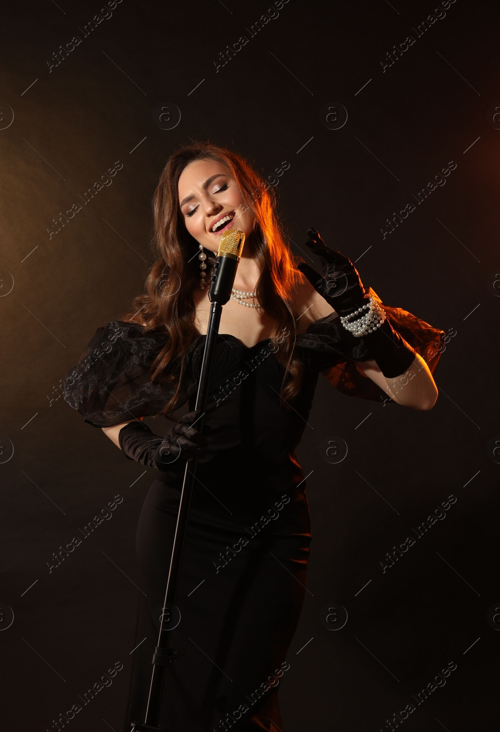 Photo of Beautiful young woman in stylish dress with microphone singing on dark background