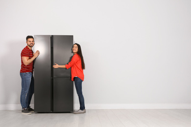 Young couple hugging refrigerator near light grey wall, space for text