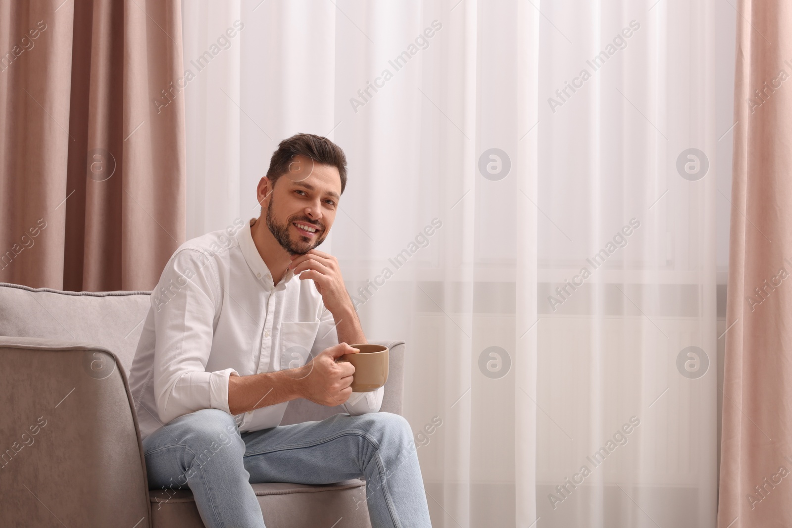 Photo of Smiling man drinking tea while resting on armchair near window with beautiful curtains at home. Space for text