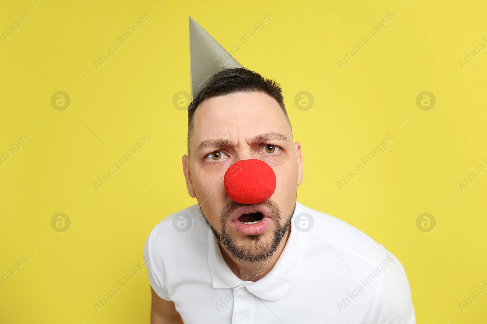 Photo of Funny man with clown nose and party hat on yellow background. April fool's day