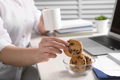Photo of Office worker with cup of drink taking chocolate chip cookie from bowl at workplace, closeup