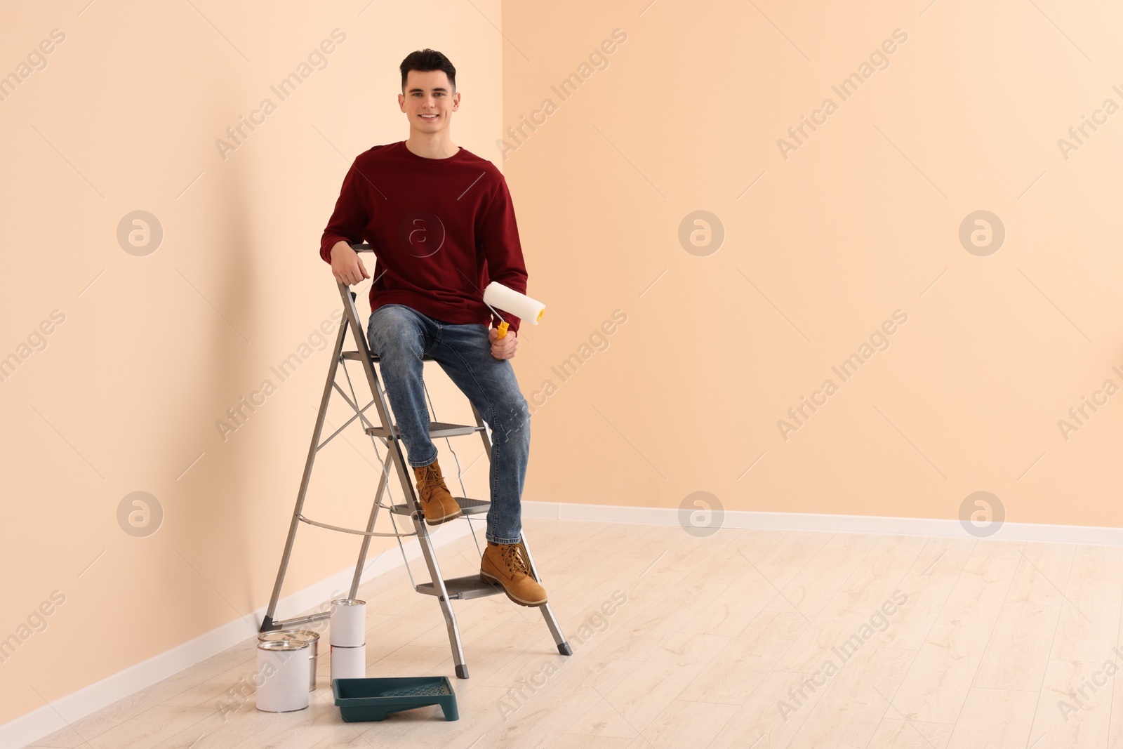 Photo of Young handsome man with roller on metal stepladder indoors, space for text. Room renovation