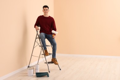 Photo of Young handsome man with roller on metal stepladder indoors, space for text. Room renovation