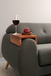 Glass of red wine and grapes on sofa with wooden armrest table in room. Interior element