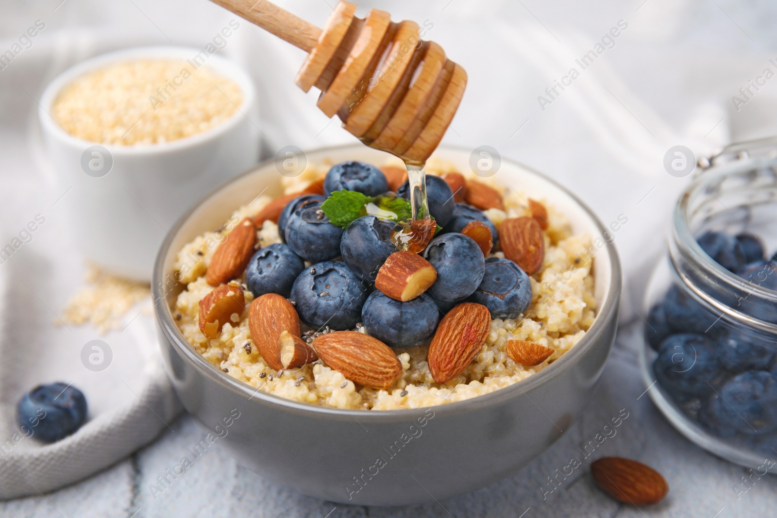 Photo of Pouring honey onto delicious cooked quinoa with almonds and blueberries in bowl on white textured table, closeup