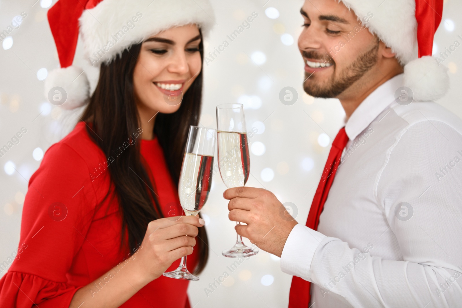 Photo of Happy young couple in Santa hats with glasses of champagne against blurred festive lights. Christmas celebration