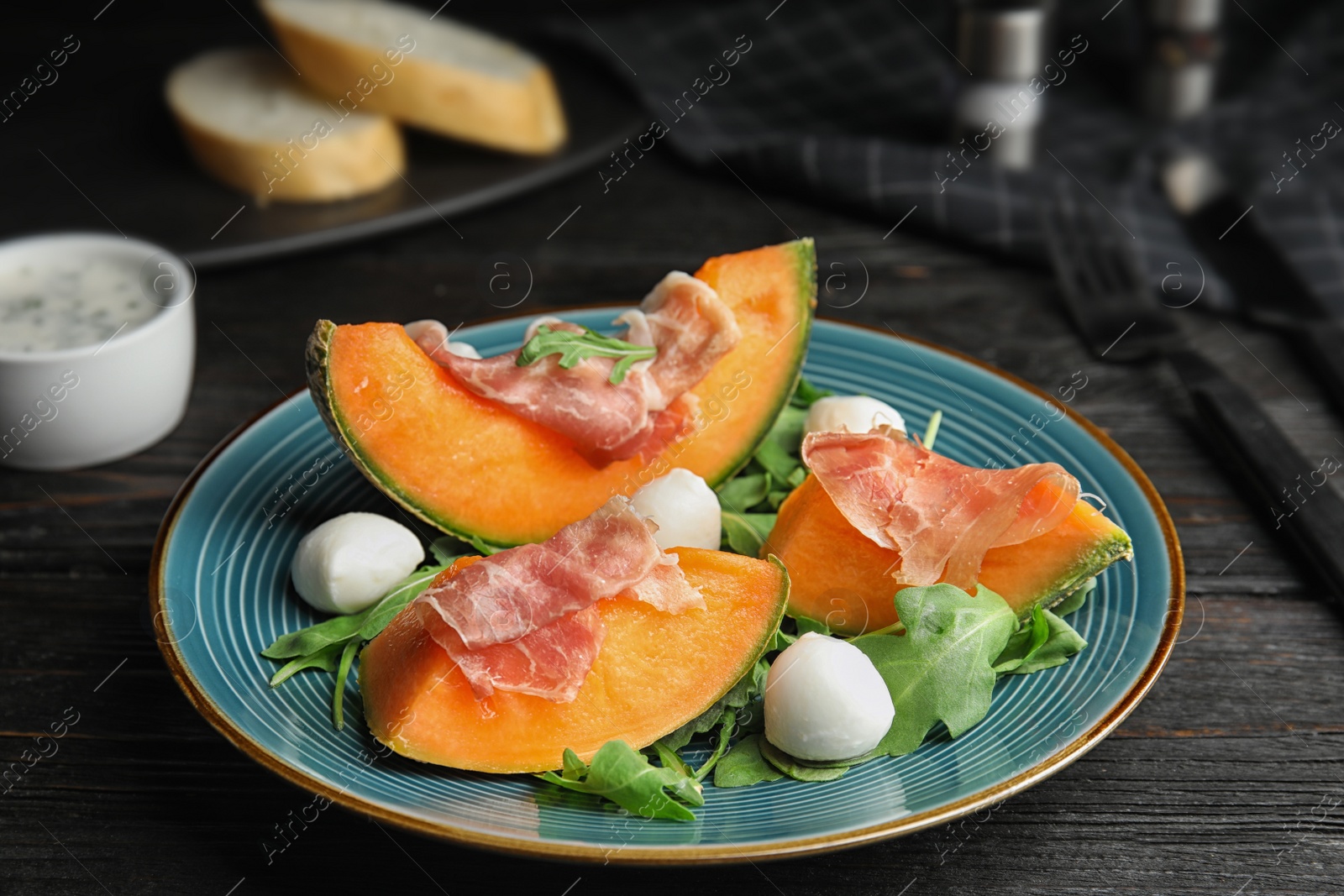 Photo of Fresh melon with prosciutto, mozzarella and arugula on black wooden table