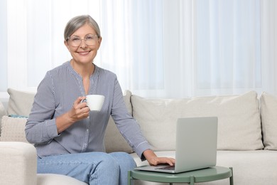 Beautiful senior woman with cup of drink using laptop at home, space for text