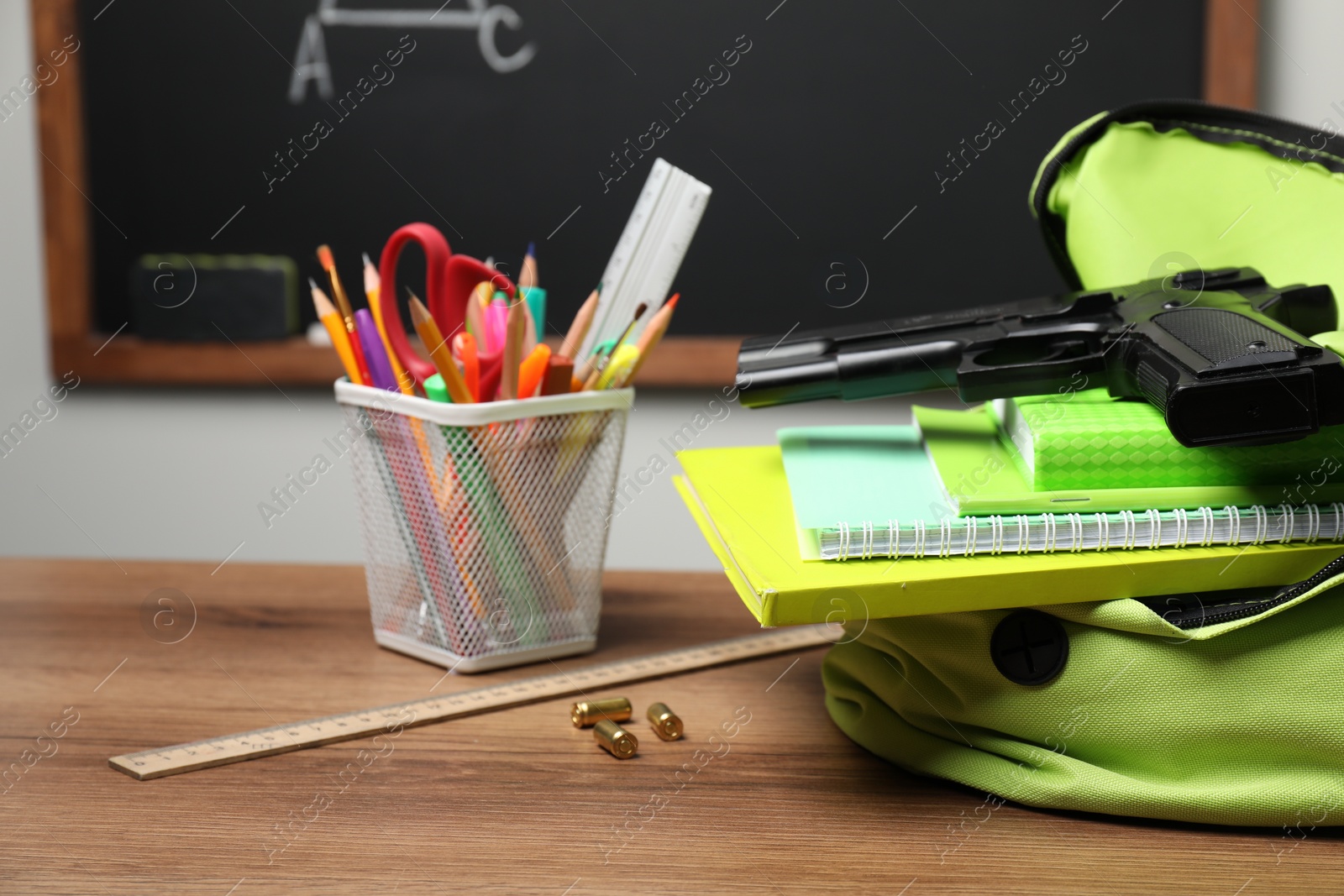 Photo of Gun, bullets and school stationery on wooden table near blackboard indoors