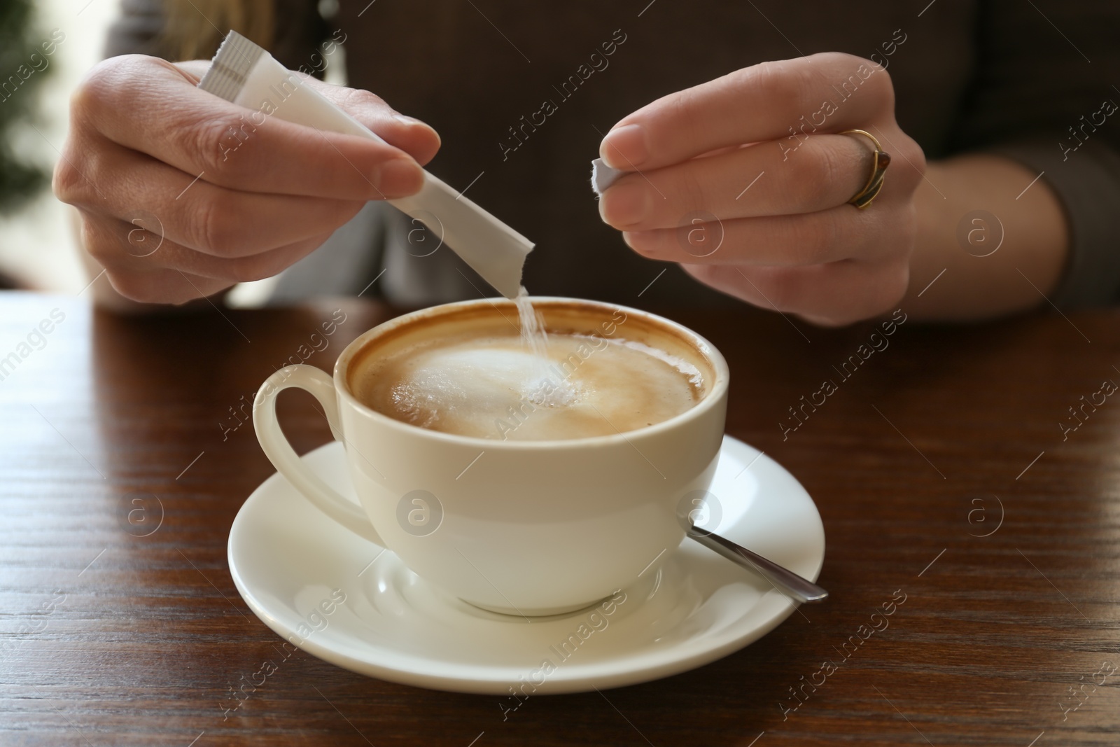 Photo of Young woman adding sugar to delicious coffee at table