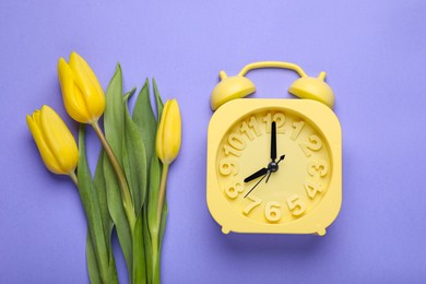 Yellow alarm clock and beautiful tulips on violet background, flat lay. Spring time