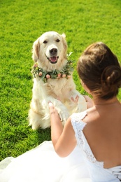 Photo of Bride and adorable Golden Retriever wearing wreath made of beautiful flowers on green grass outdoors, closeup