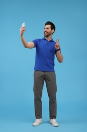 Smiling man taking selfie with smartphone and showing peace sign on light blue background