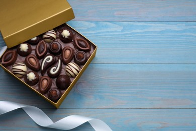 Open box of delicious chocolate candies and color ribbon on light blue wooden table, flat lay. Space for text