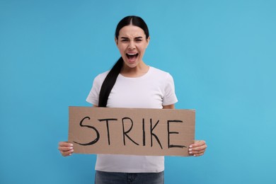 Angry woman holding cardboard banner with word Strike on light blue background