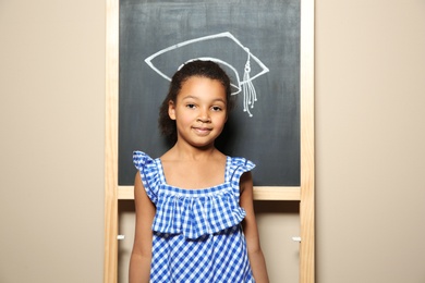 African-American child standing at blackboard with chalk drawn academic cap. Education concept