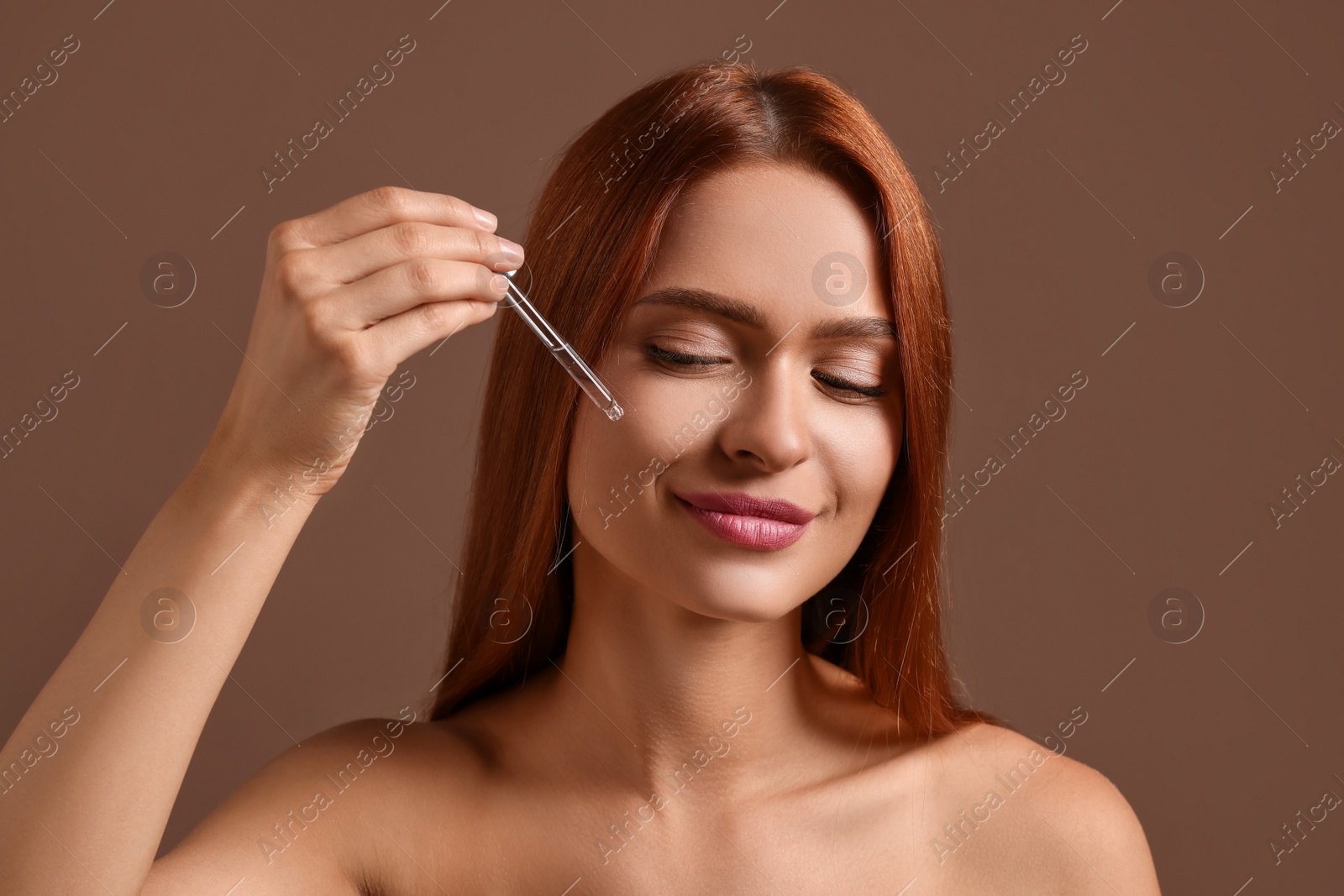 Photo of Beautiful young woman applying cosmetic serum onto her face on brown background