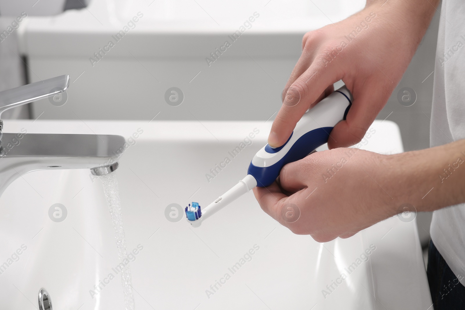 Photo of Man holding electric toothbrush near flowing water above sink in bathroom, closeup