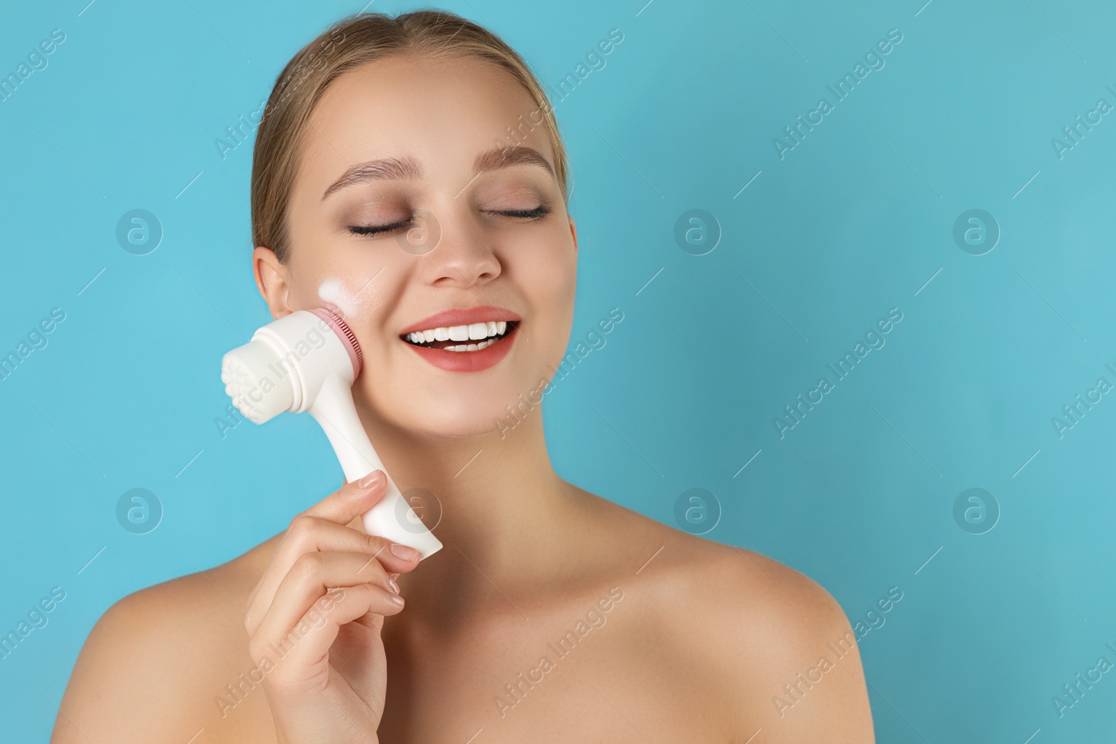 Photo of Young woman washing face with brush and cleansing foam on light blue background. Cosmetic products