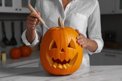 Photo of Woman carving pumpkin for Halloween at white marble table in kitchen, closeup