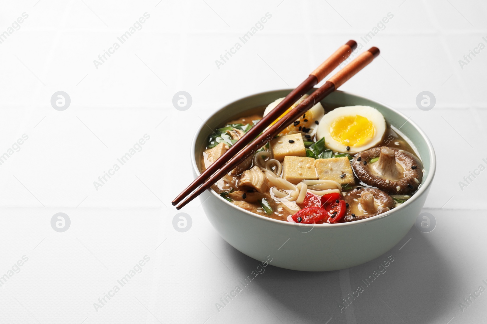 Photo of Noodle soup. Bowl of delicious ramen and chopsticks on white tiled table, space for text