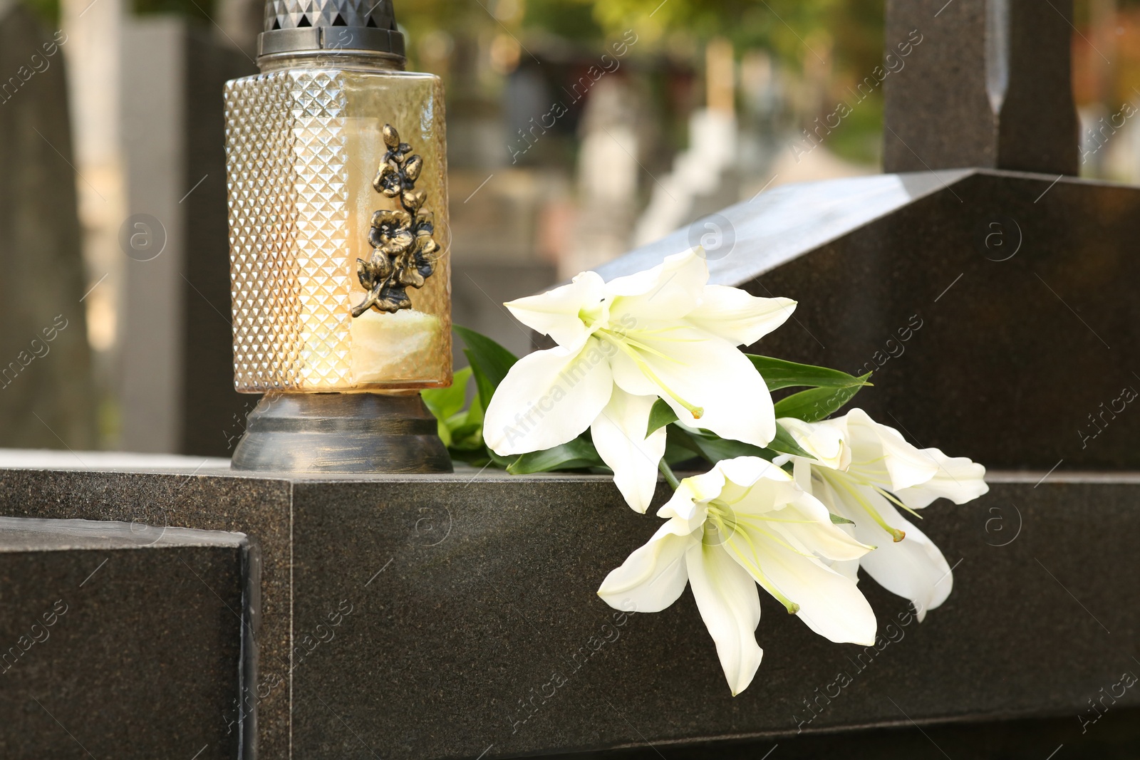 Photo of White lilies and grave light on grey granite tombstone outdoors. Funeral ceremony