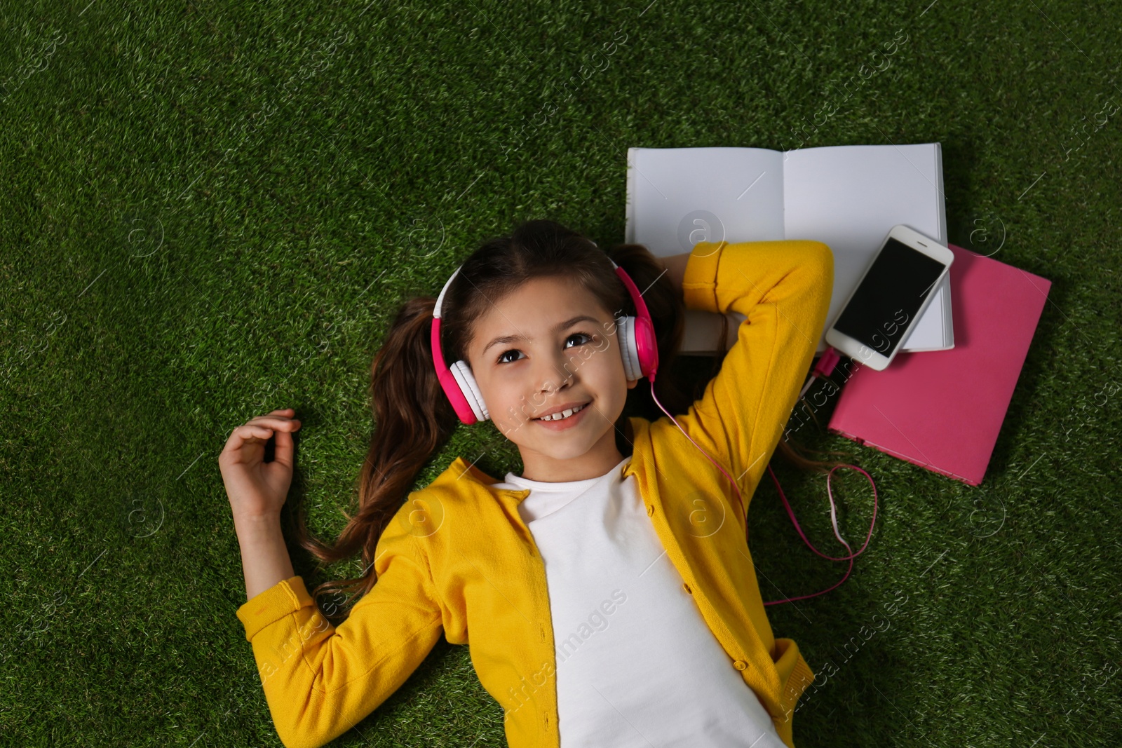 Photo of Cute little girl listening to audiobook on grass, top view