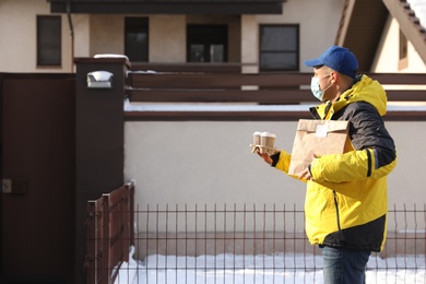 Photo of Courier in medical mask holding takeaway food and drinks near house outdoors. Delivery service during quarantine due to Covid-19 outbreak