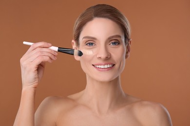Woman applying foundation on face with brush against brown background