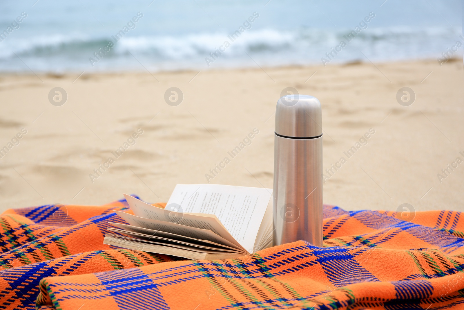 Photo of Metallic thermos with hot drink, open book and plaid on sandy beach near sea