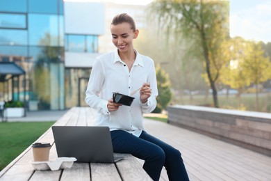 Smiling businesswoman with lunch box sitting near laptop and paper cup outdoors. Space for text