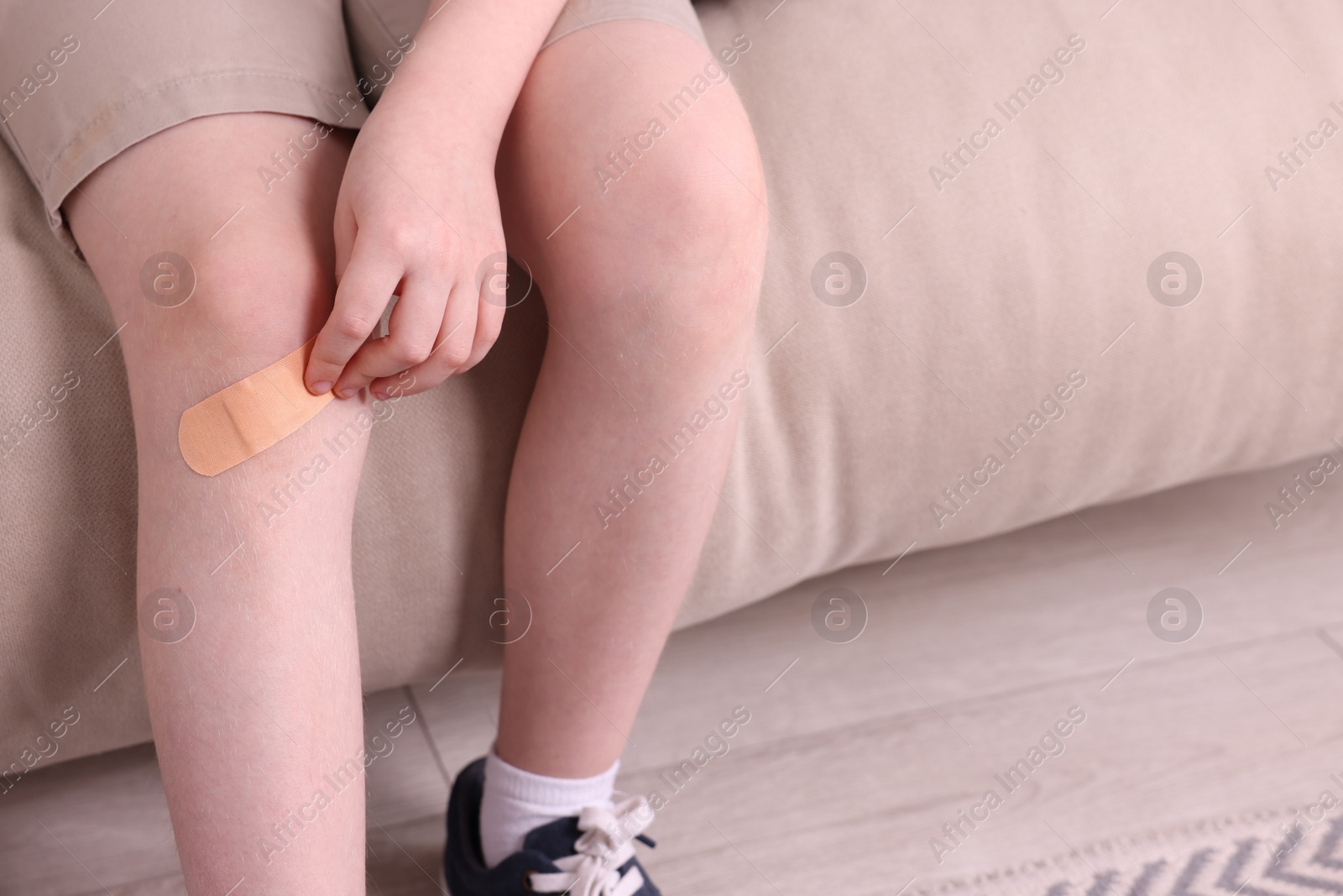 Photo of Little boy putting sticking plaster onto knee on sofa, closeup. Space for text