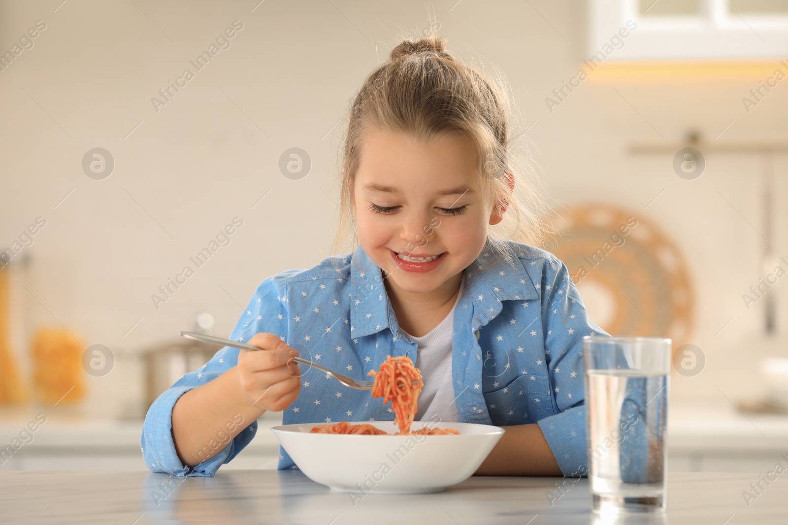 Photo of Cute little girl eating tasty pasta at table in kitchen