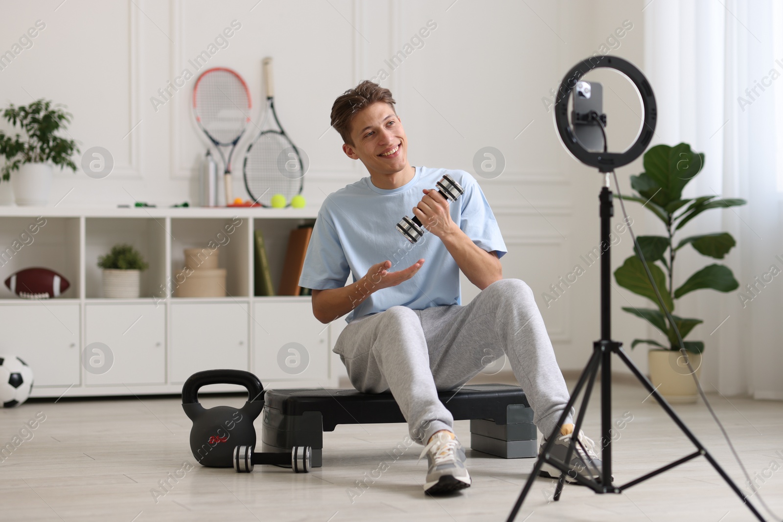 Photo of Smiling sports blogger holding dumbbell while streaming online fitness lesson with smartphone at home