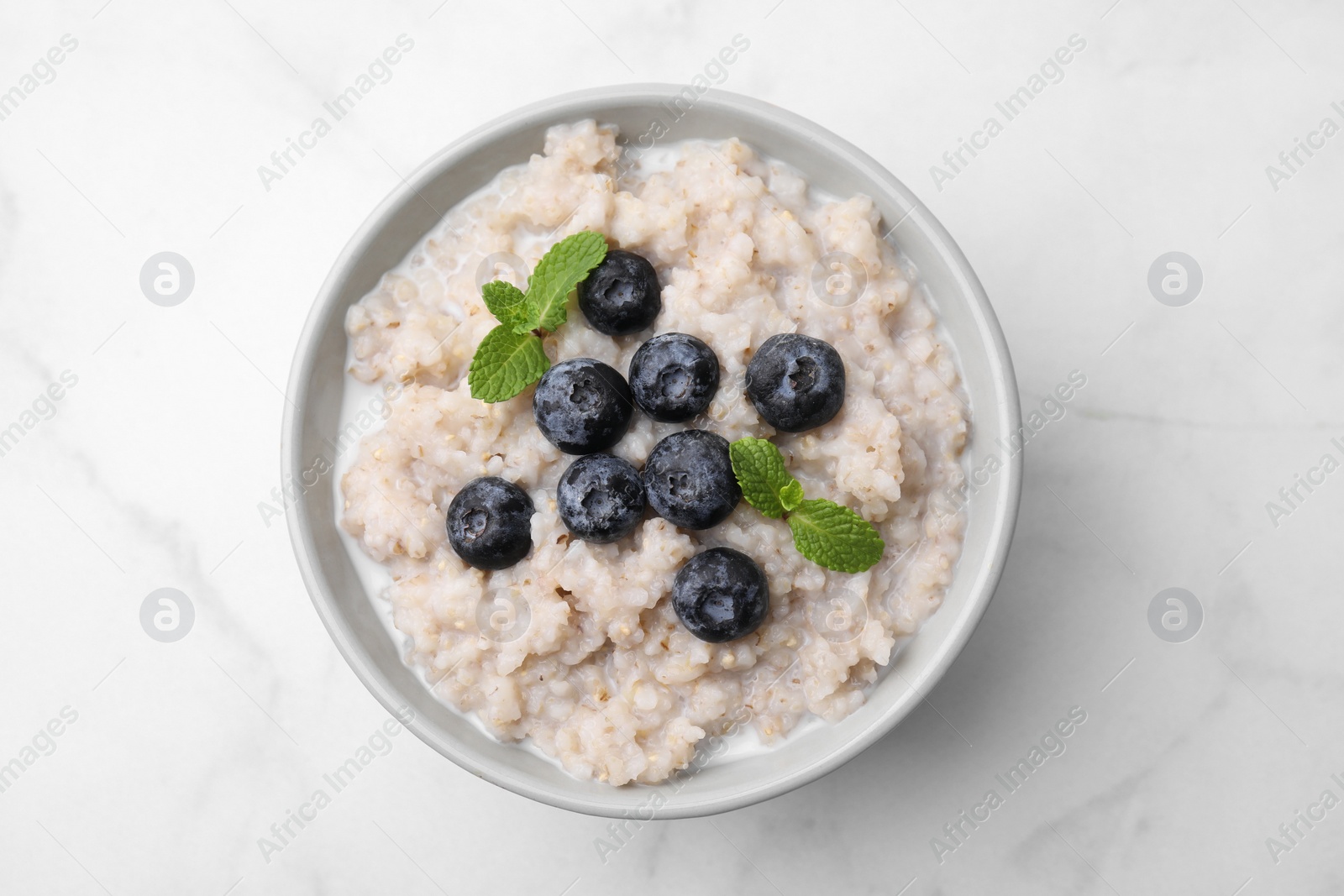 Photo of Delicious barley porridge with blueberries and mint in bowl on white marble table, top view