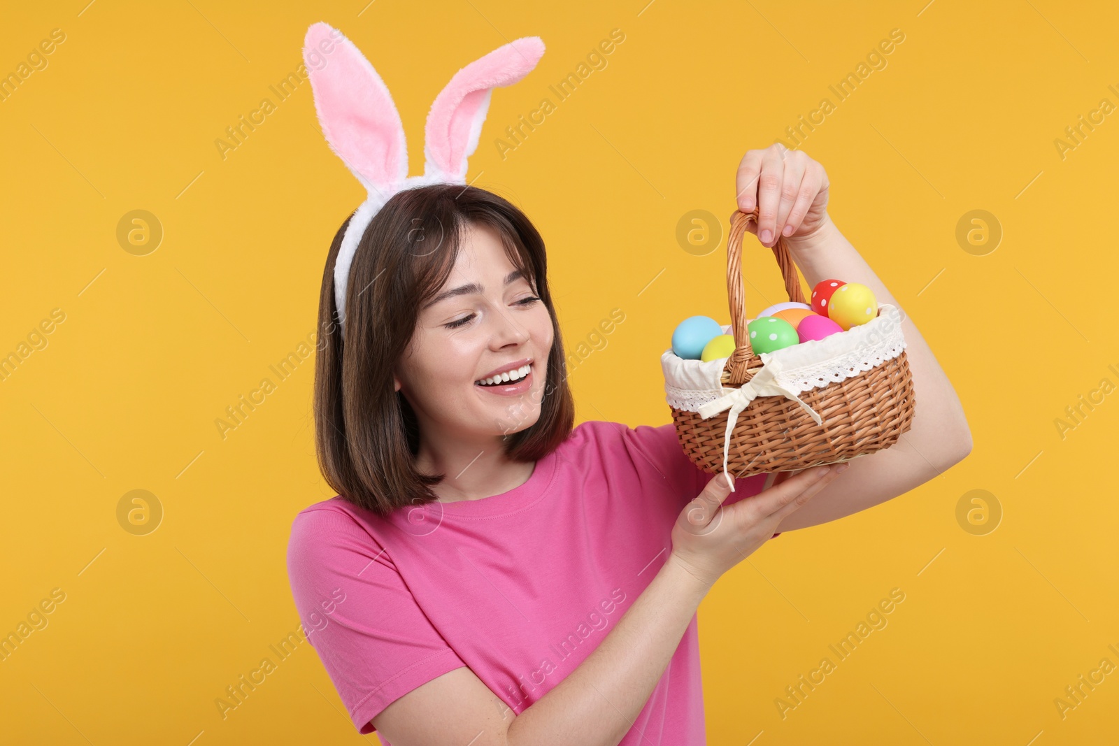 Photo of Easter celebration. Happy woman with bunny ears and wicker basket full of painted eggs on orange background
