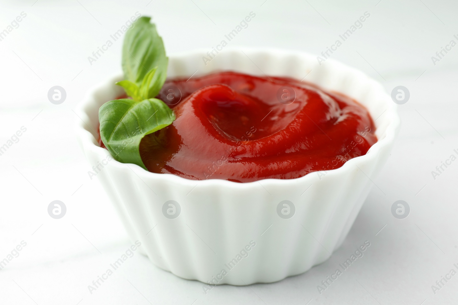 Photo of Bowl of tasty ketchup and basil on white marble table, closeup