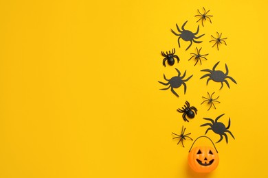 Flat lay composition with plastic pumpkin basket and spiders on yellow background, space for text. Halloween celebration