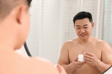 Photo of Handsome man with jar of body cream near mirror indoors