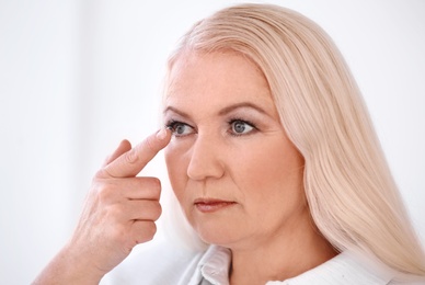 Senior woman putting contact lens in her eye on light background