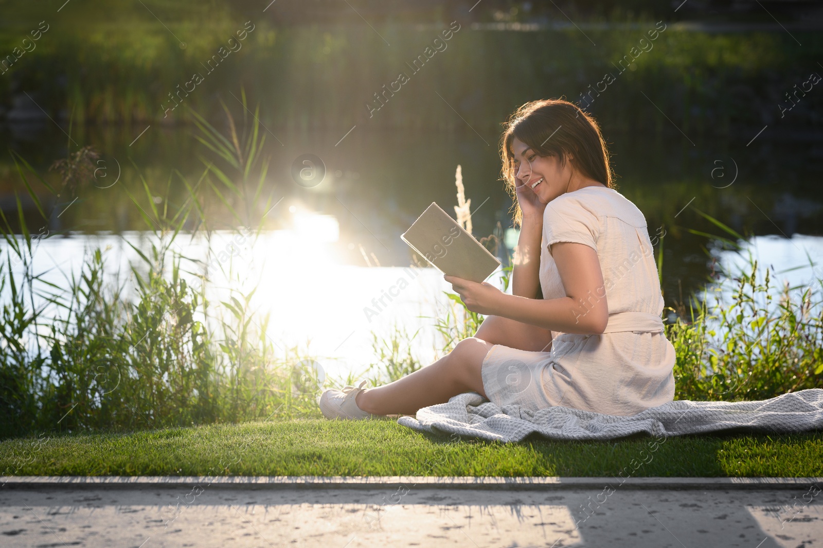 Photo of Young woman reading book near lake on sunny day