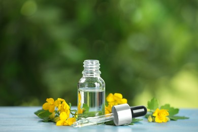 Photo of Bottle of natural celandine oil near flowers on light blue wooden table outdoors