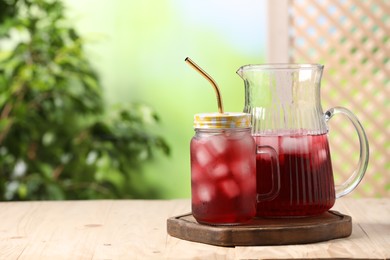 Photo of Refreshing hibiscus tea with ice cubes on wooden table against blurred green background. Space for text