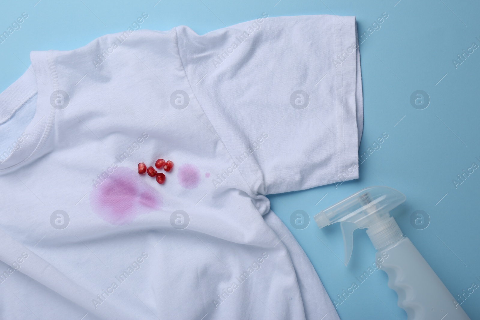 Photo of White shirt with fruit juicy stains, detergent and pomegranate seeds on light blue background, flat lay