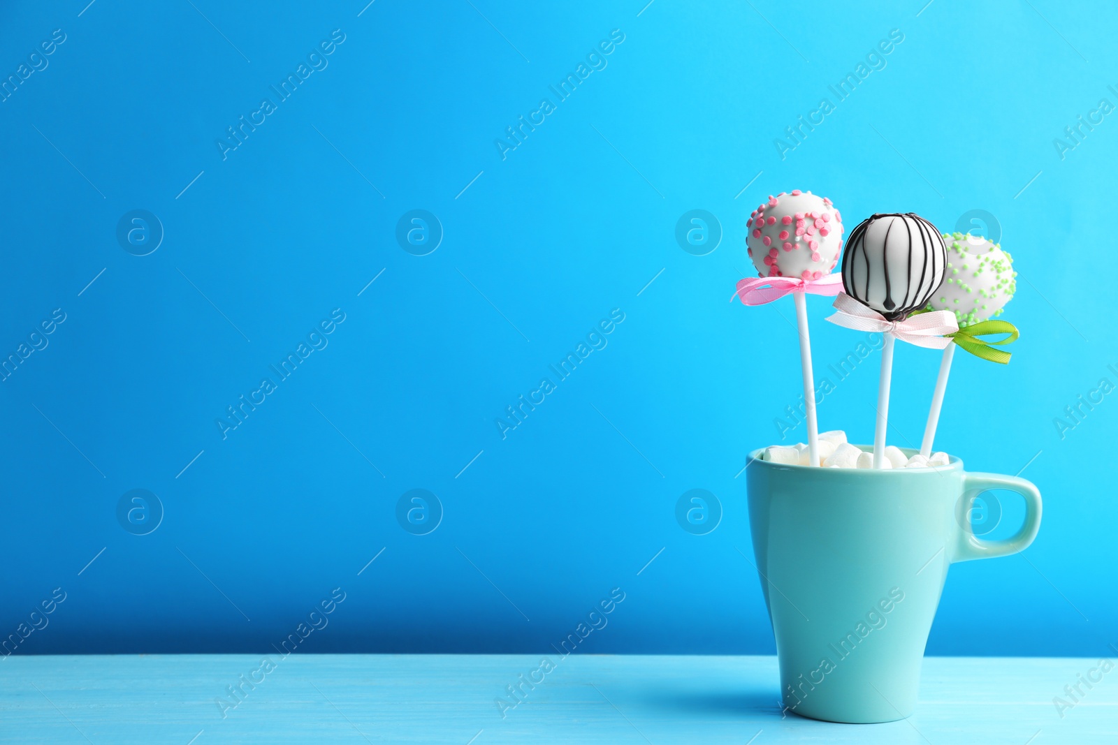 Photo of Tasty cake pops in cup with marshmallow on wooden table against light blue background. Space for text