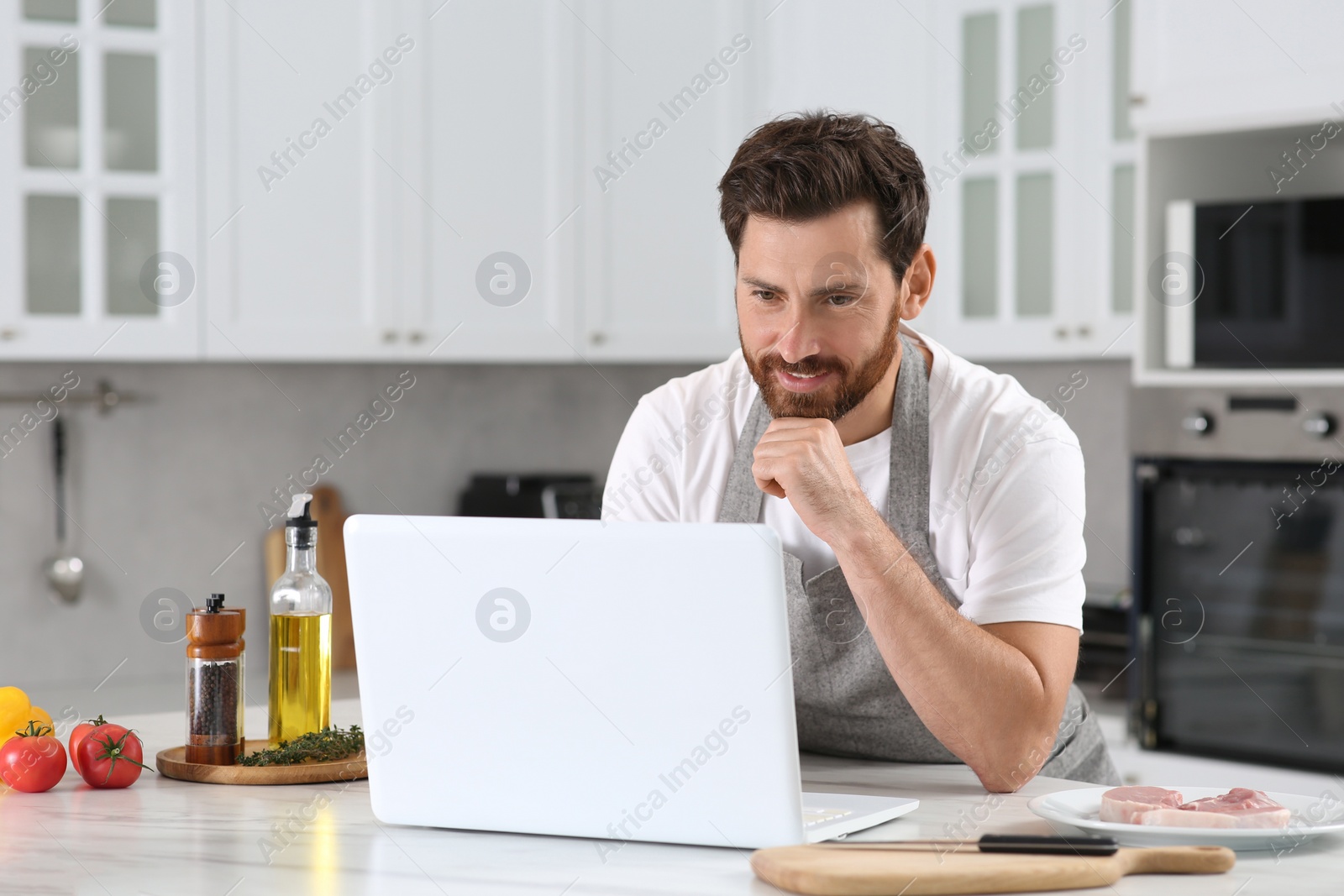 Photo of Man making dinner while watching online cooking course via laptop in kitchen